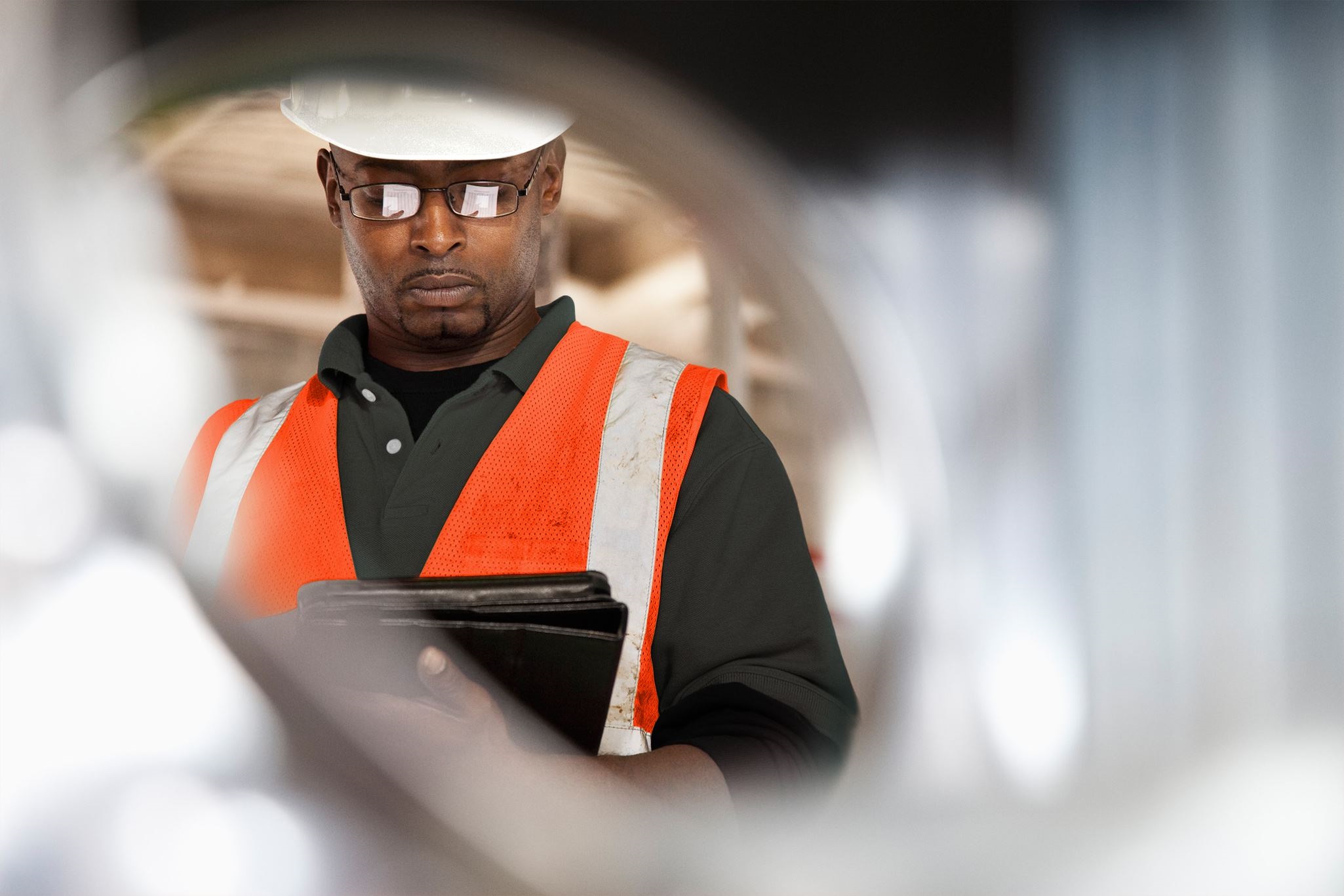 A worker in an orange hi-vis vest and hard hat holds a clipboard, observed through a circular opening in a construction setting