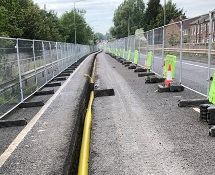 Road construction site with a narrow trench containing a yellow utility pipe, fenced off on both sides with metal barriers and green safety signs.