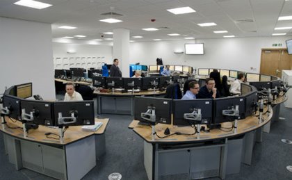 A spacious control room with curved desks, multiple computer monitors, and focused staff. The atmosphere is busy and professional, displaying teamwork and technology.