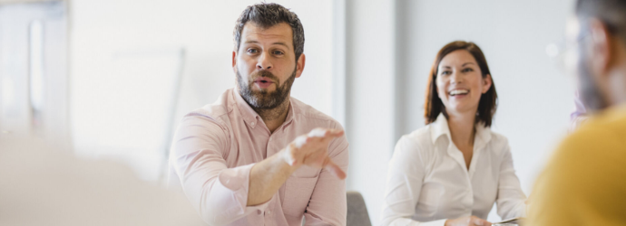 A group of professionals engage in a discussion during a meeting, with one participant actively gesturing while speaking.
