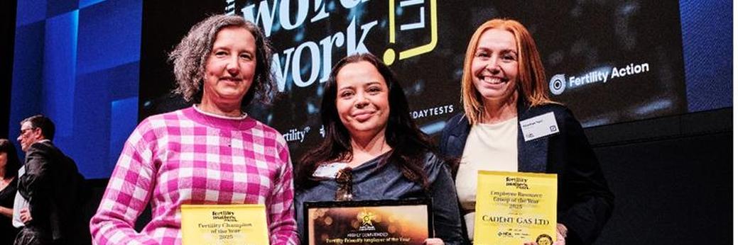 Three women standing on stage holding award certificates and plaques, smiling at the camera. A large screen behind them displays event branding related to Fertility at Work and Fertility Action.