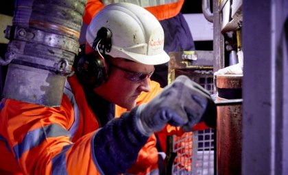 A worker in protective gear, including helmet and ear protection, operates machinery with focus. The scene is lit by artificial light, creating an industrial ambiance.