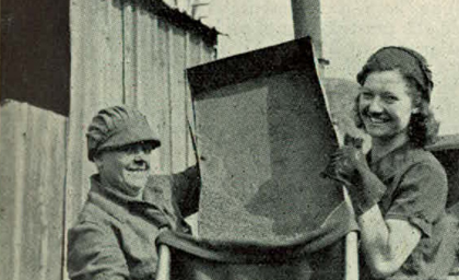 Two women in work attire, wearing caps and gloves, smile while holding a large tray outside a wooden building. The tone is positive and industrious.