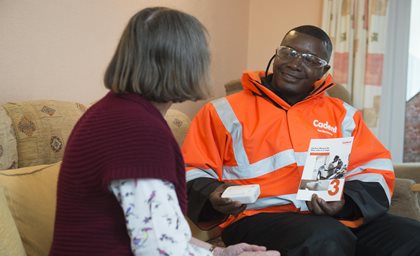 A man in an orange safety jacket sits on a sofa, smiling and holding a brochure and device, talking to a woman with grey hair in a cozy setting.