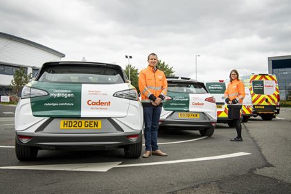 Two cars with Cadent branding for hydrogen and electricity parked in a lot, with two people in orange safety jackets standing nearby and a Cadent service van in the background.