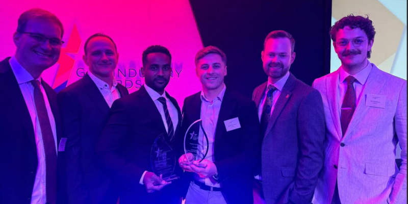 Six men in formal business attire standing together at an awards event, two of them holding glass trophies. The group is lit with strong pink and purple stage lighting, with an awards backdrop visible behind them.