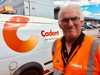 Man in an orange Cadent vest smiles in front of a company van with a logo. The scene is bright and outdoor, conveying a friendly, professional tone.