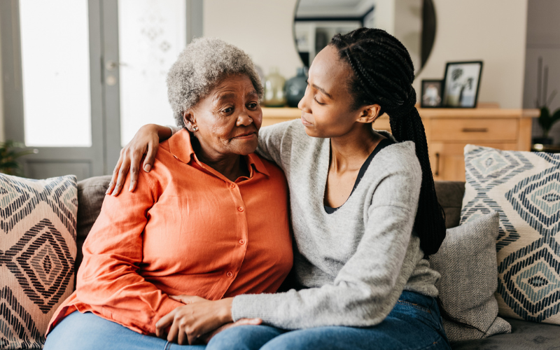 Younger woman supporting and comforting an elderly woman while sitting on a sofa.
