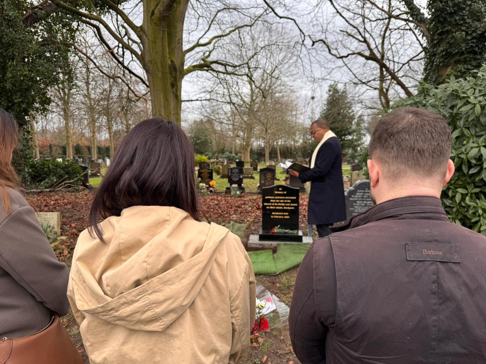 People attending a graveside service in a cemetery, standing before a headstone while an officiant reads during a memorial ceremony on an overcast day.