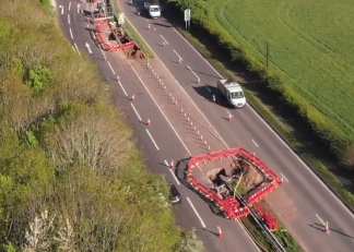 Roadworks on a dual carriageway with red barriers and traffic cones around excavation sites, vehicles passing by, and green fields on one side.