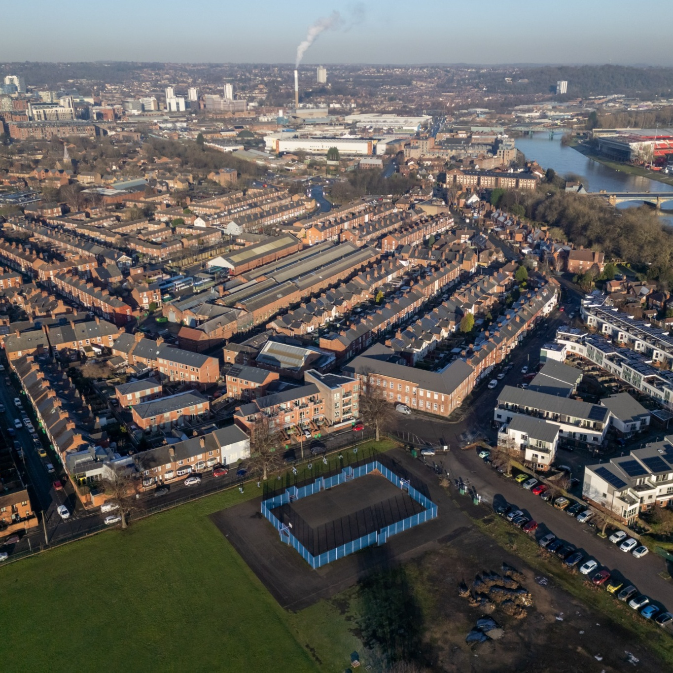 Aerial view of a residential area with rows of red-brick houses, intersecting roads, a sports court, and a smokestack in the distance under a clear sky.