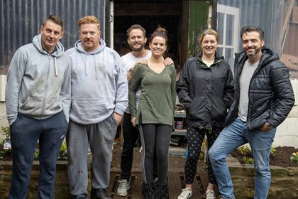 Six people standing together in front of a rustic building with corrugated metal walls and a door.