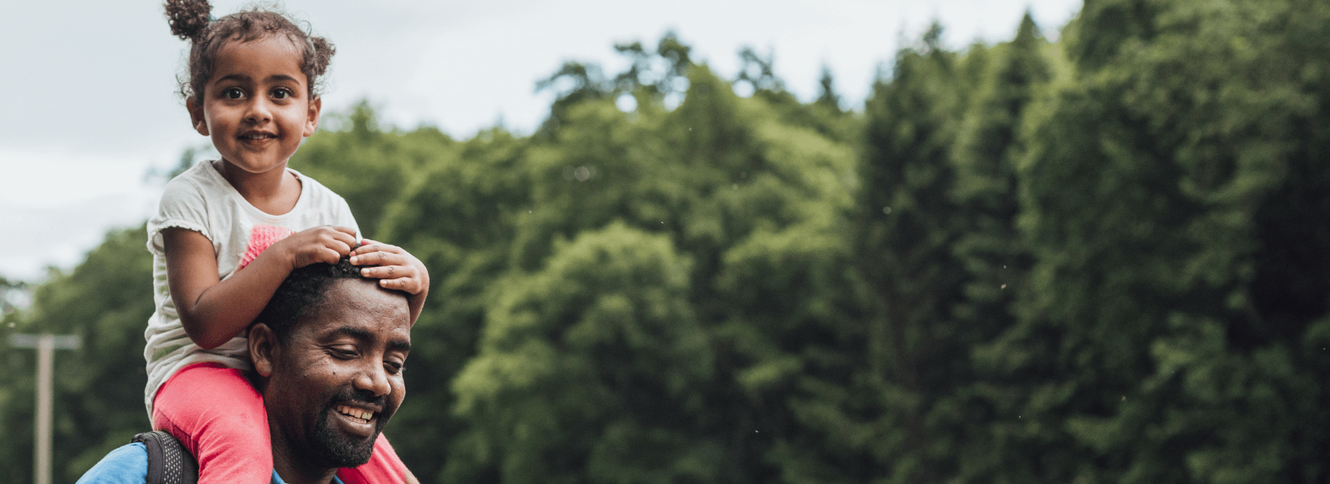 Father carrying his young daughter on his shoulders whilst on a walk through a park with wooded area in the background