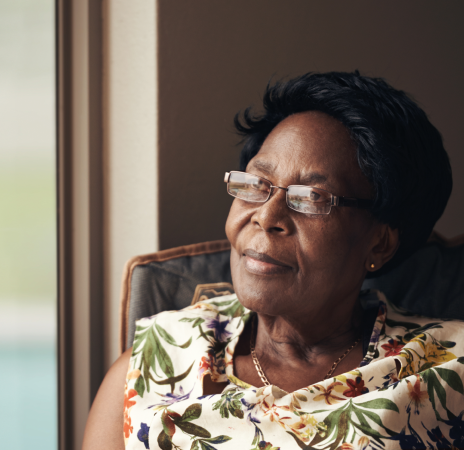 A woman with short black hair sits contemplatively in a cozy chair, dressed in a floral-patterned blouse, with warm sunlight illuminating her surroundings.