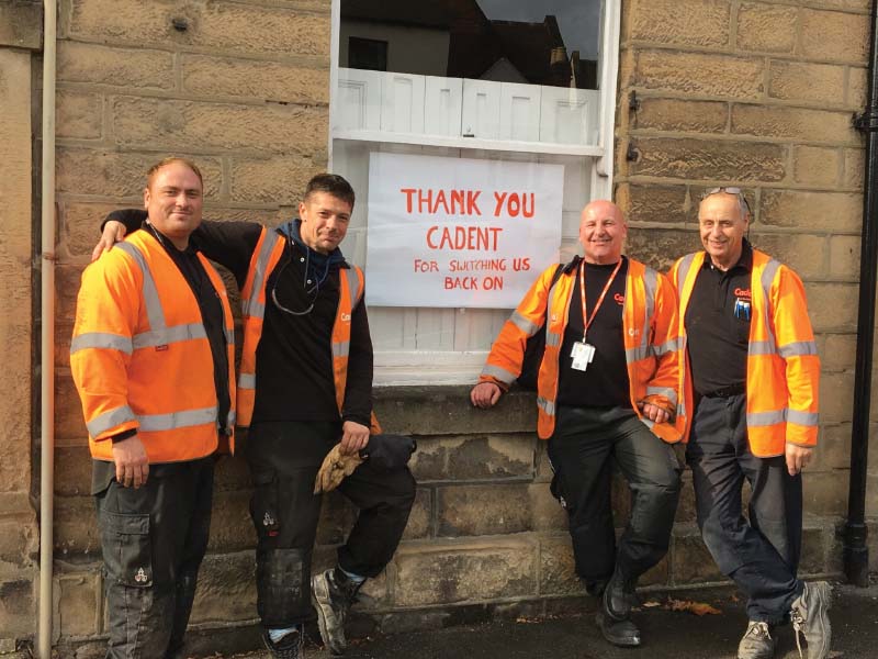 Four workers in hi-vis orange jackets stand in front of a window with a sign thanking Cadent for their service.