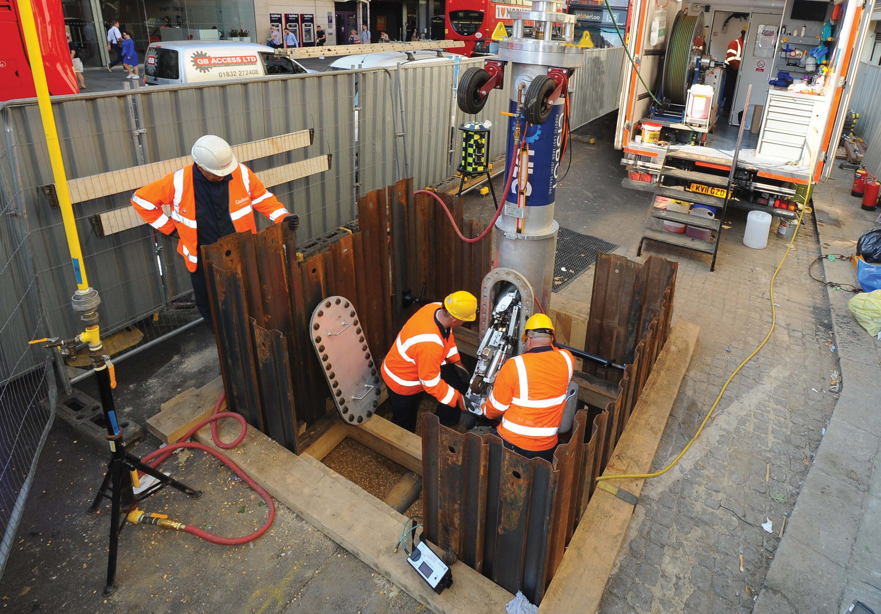 Three construction workers in safety gear operate machinery in a deep trench surrounded by steel sheeting, with equipment visible nearby.