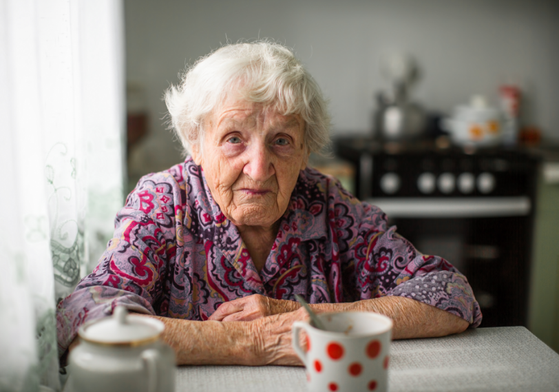 Elderly lady, dressed in a floral shirt, looking seriously into the camera - sat at the kitchen table drinking a cup of tea