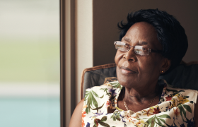 A woman with short black hair sits contemplatively in a cozy chair, dressed in a floral-patterned blouse, with warm sunlight illuminating her surroundings.