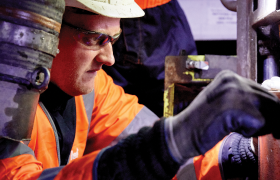 A worker in an orange safety jacket and helmet inspects machinery closely. He wears safety glasses and gloves, concentrating on his task in an industrial setting.