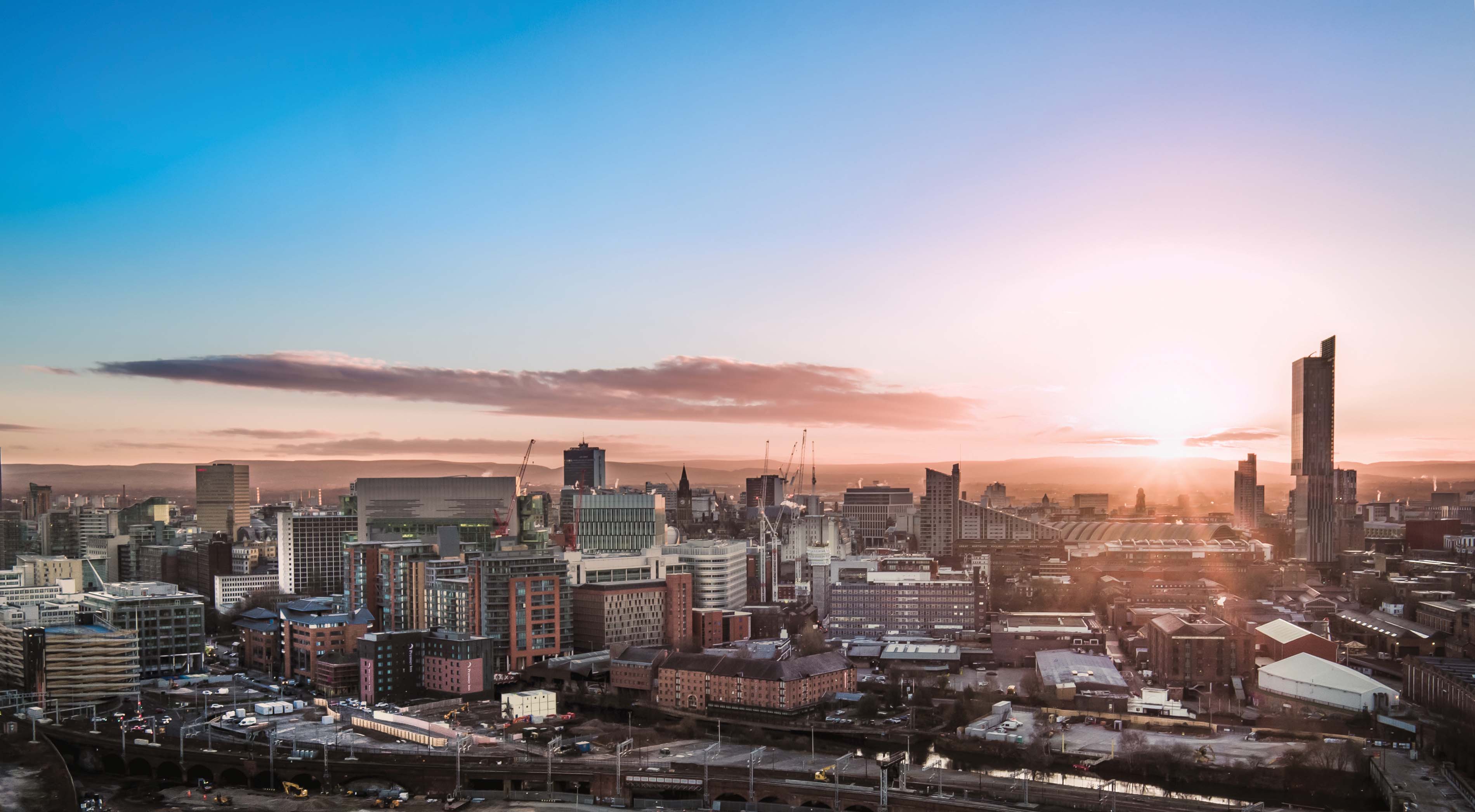 A panoramic view of Manchester at sunset, showcasing a skyline of skyscrapers and low-rise buildings against a vibrant sky.