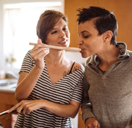 Two people in a kitchen enjoy cooking together. One person holds a wooden spoon, offering a taste to the other, who smiles. Sunlight filters through a window.