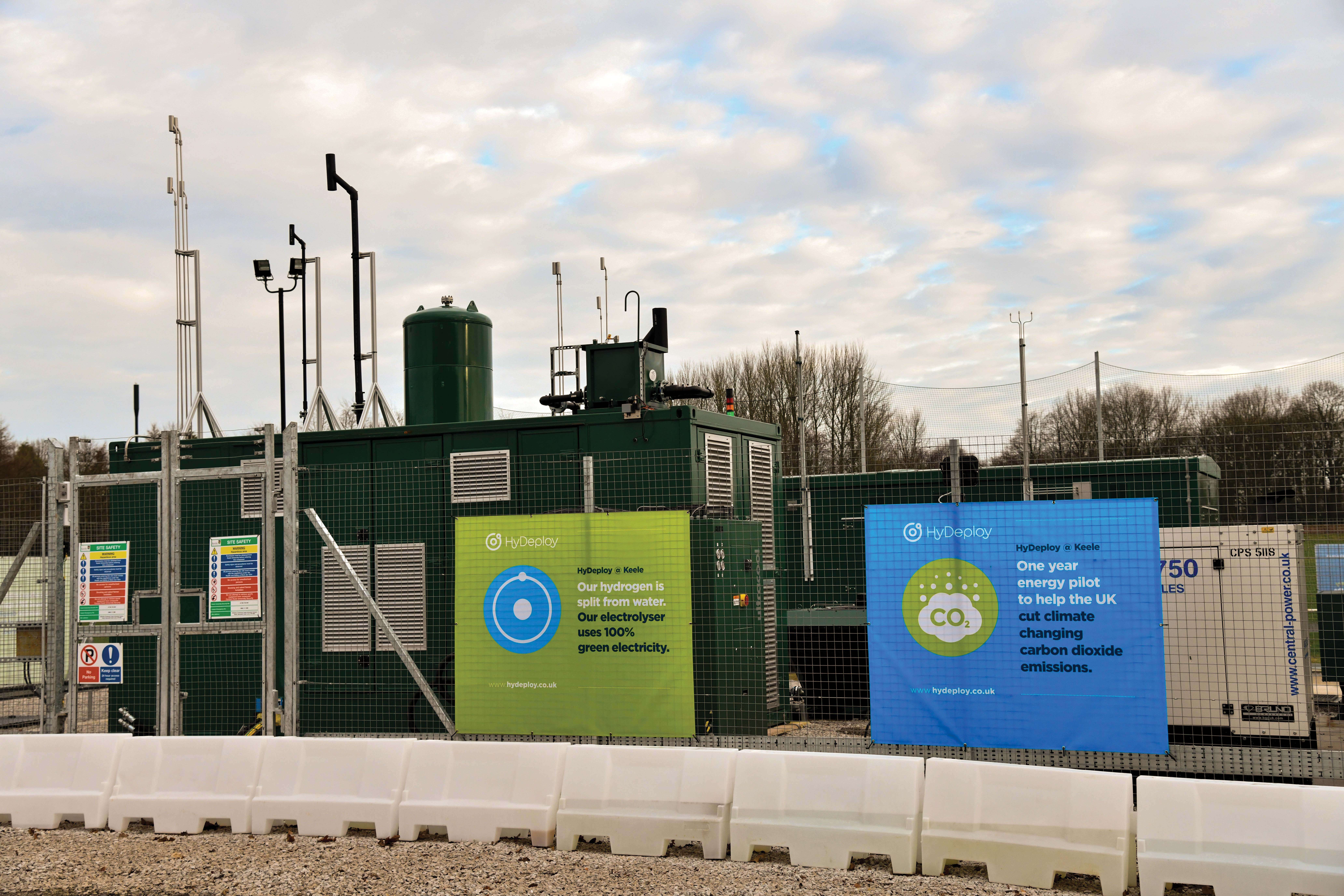 A green energy facility with banners promoting hydrogen production from water and carbon dioxide reduction, surrounded by fencing and barriers.