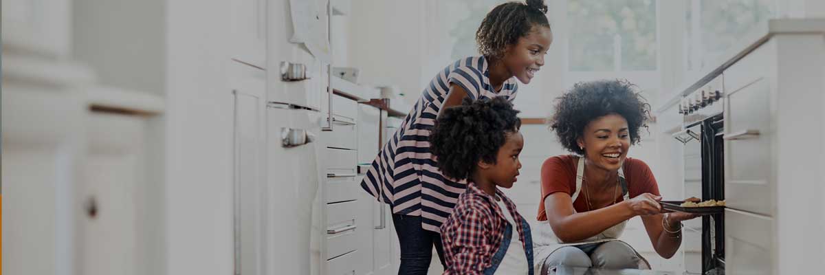 A woman and two children gather around an oven in a bright kitchen, eagerly watching as food is being removed from the oven