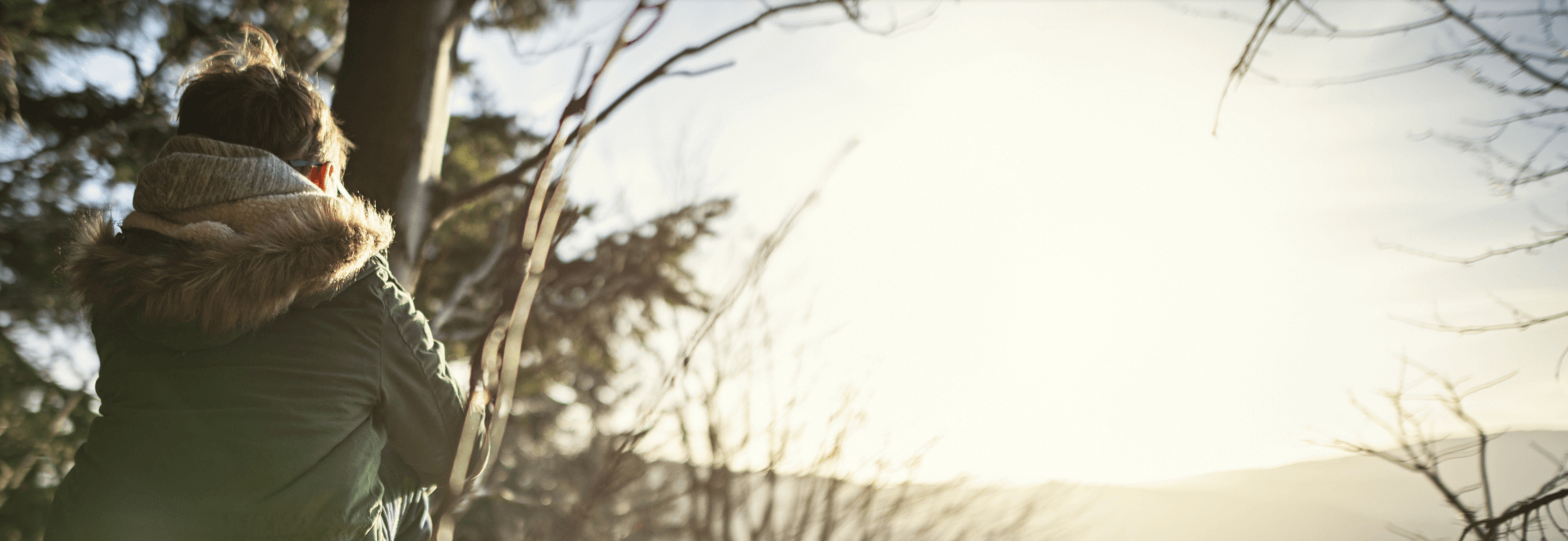 Young boy outside looking out at a wooded landscape whilst the sun sets, dressed in a green coat with fur hood