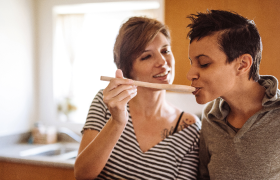 Two people in a kitchen enjoy cooking together. One person holds a wooden spoon, offering a taste to the other, who smiles. Sunlight filters through a window.