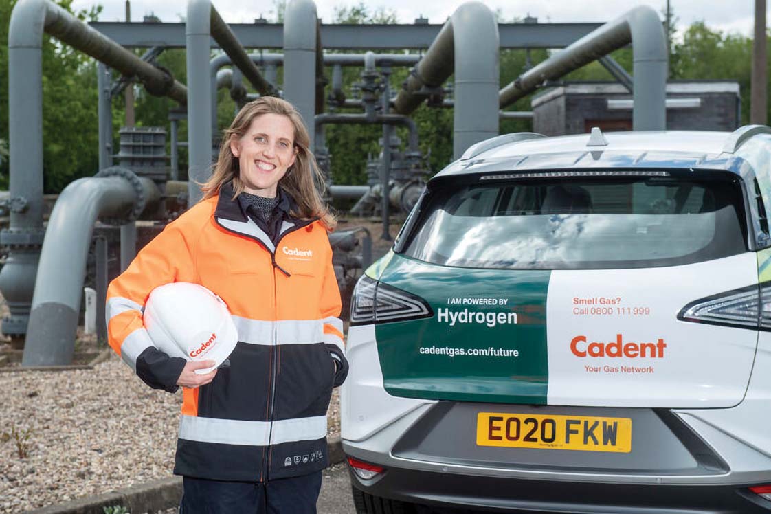 A Cadent employee in an orange and black jacket stands with a hard hat, next to a hydrogen-powered vehicle in an industrial setting.