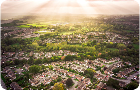 Aerial view of a residential suburban area with houses, roads, and greenery, under a sunset sky. A railway track cuts across the neighbourhood.