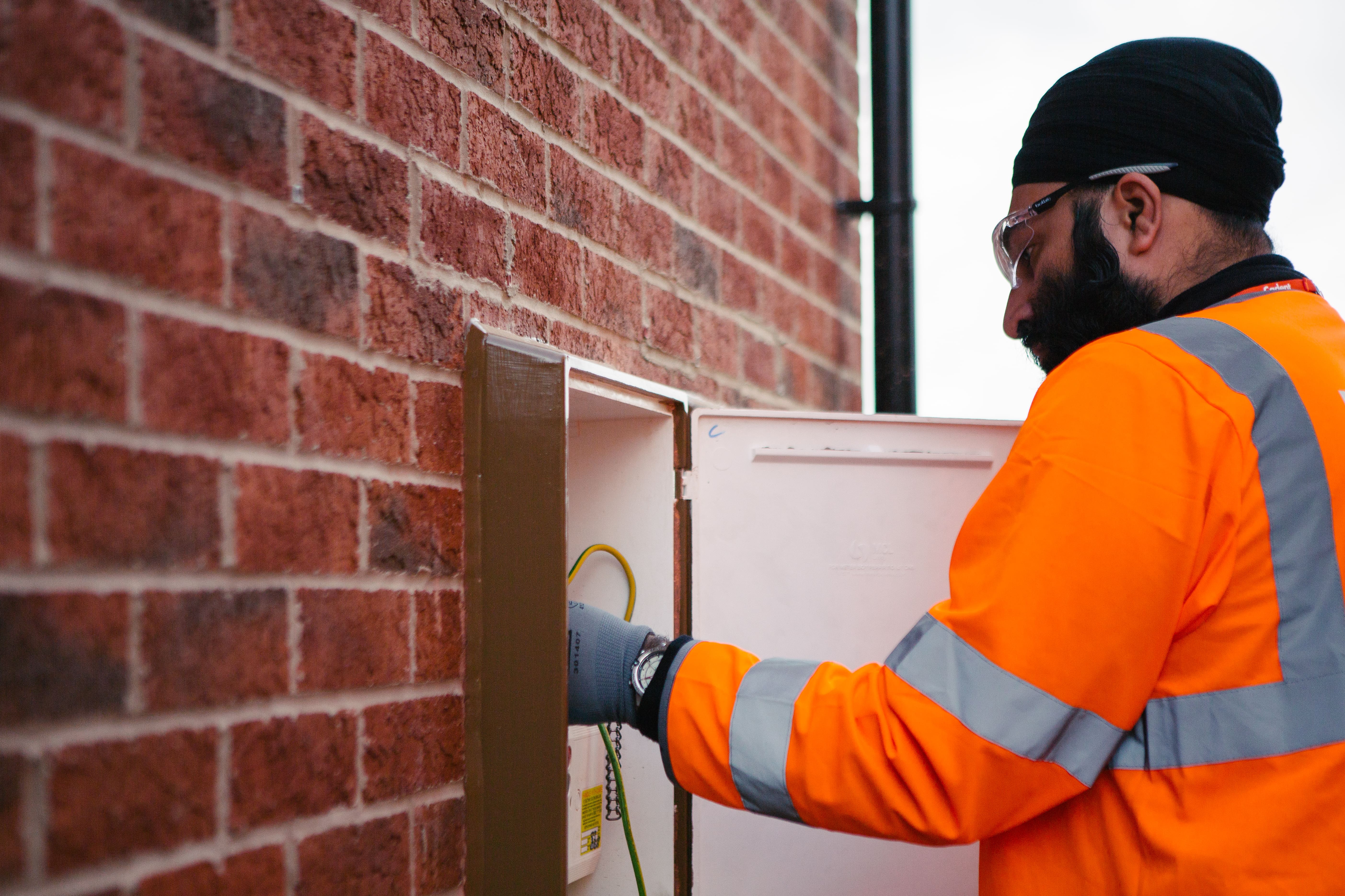 A Cadent engineer in an orange hi-vis jacket is inspecting a utility box on a brick wall, with tools and wires visible inside.