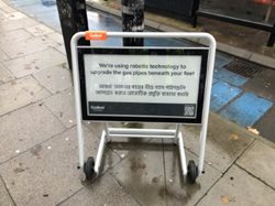 A sign on pavement announcing the use of robotics to upgrade underground pipelines, with text in English and multiple languages.