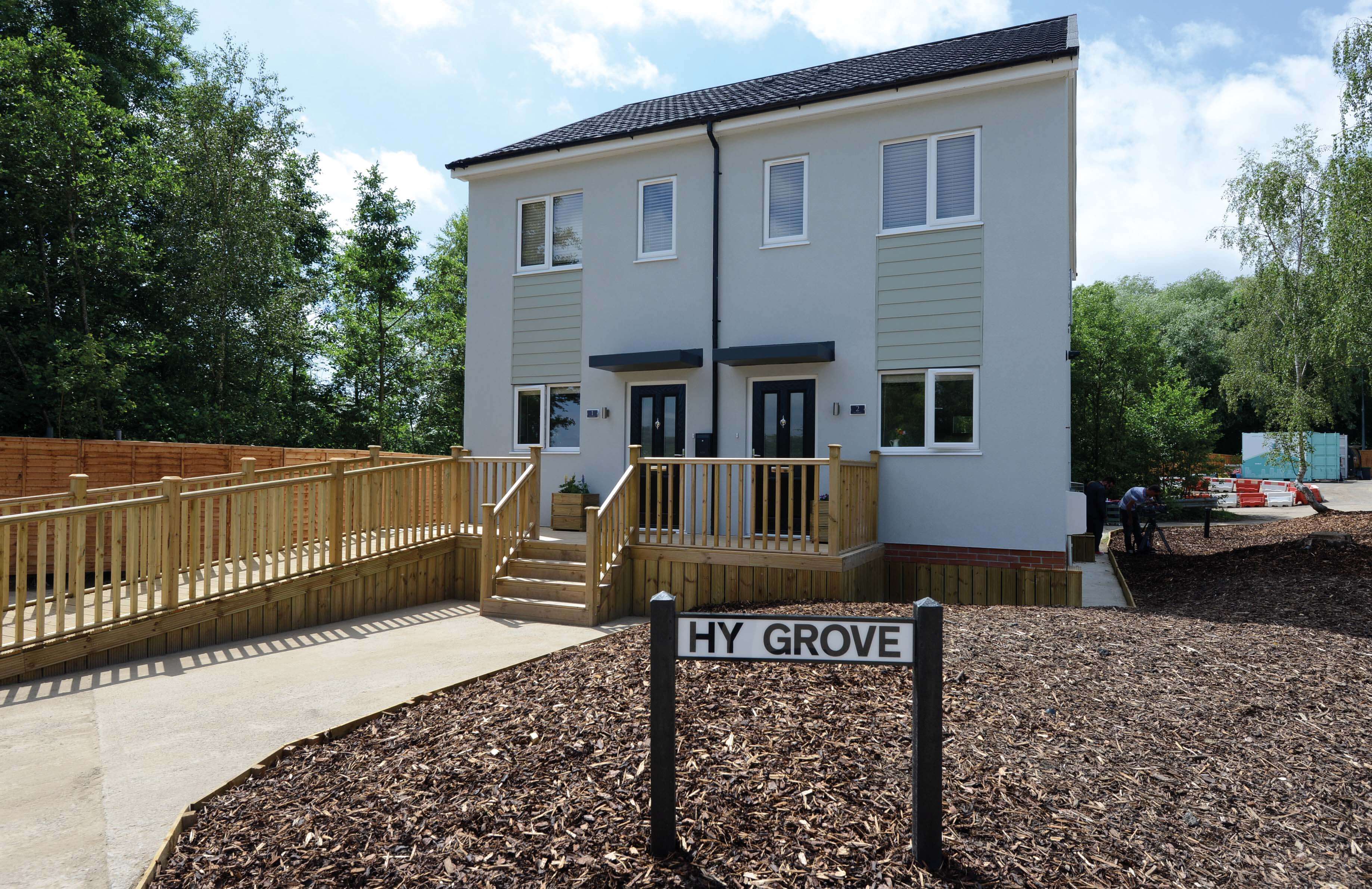 A modern two-story house with a wooden deck, surrounded by greenery, features a sign stating 