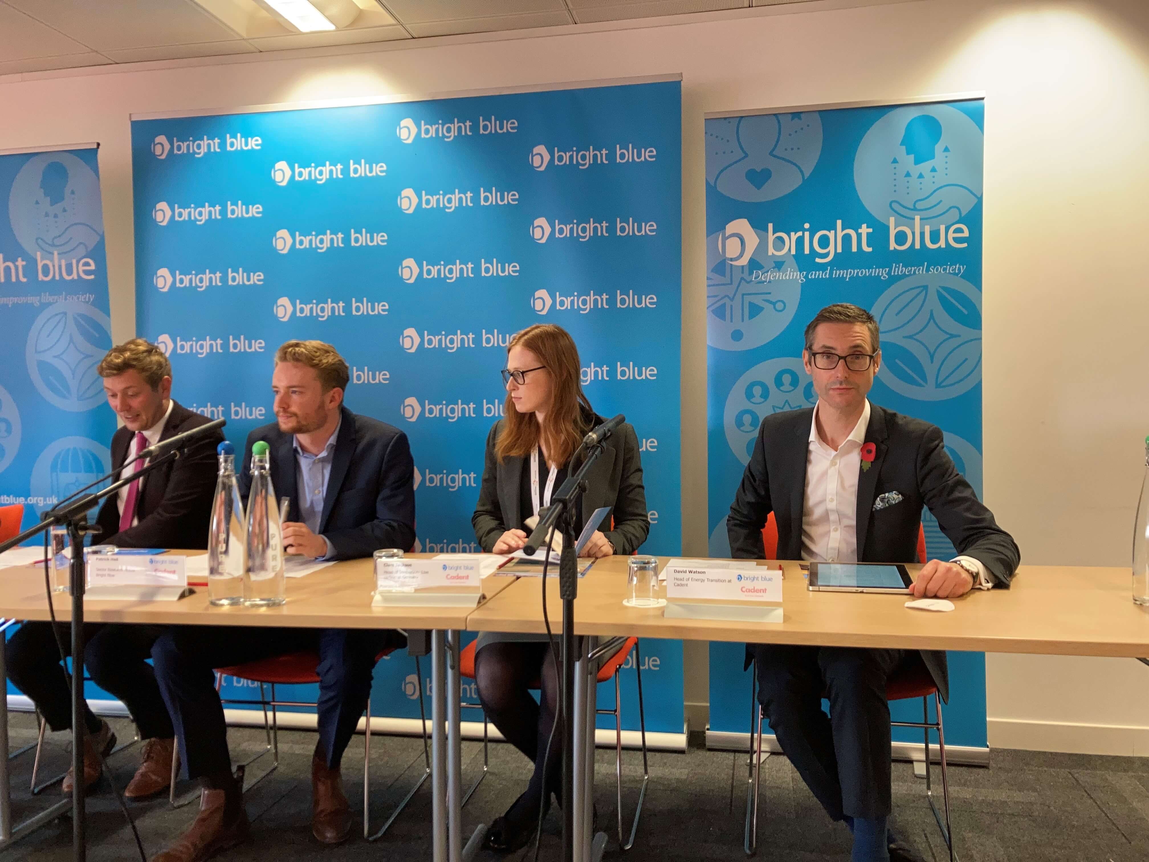 A panel of speakers discusses topics at a conference, seated at a table with water bottles and microphones against a bright blue branded background.