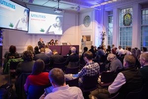 A conference room filled with attendees, focused on a presentation about utilities, with speakers at the front and screens displaying slides.