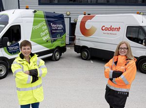 Two workers in safety gear stand confidently in front of electric and gas service vehicles parked side by side, showcasing their companies.