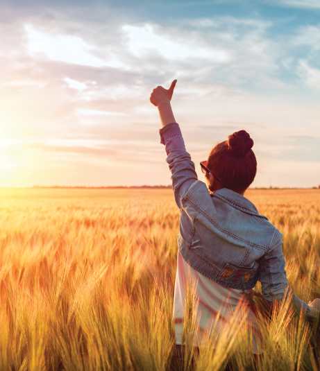A woman wearing a denim jacket stands in a golden wheat field, joyfully raising her hand toward a colourful sunset sky.