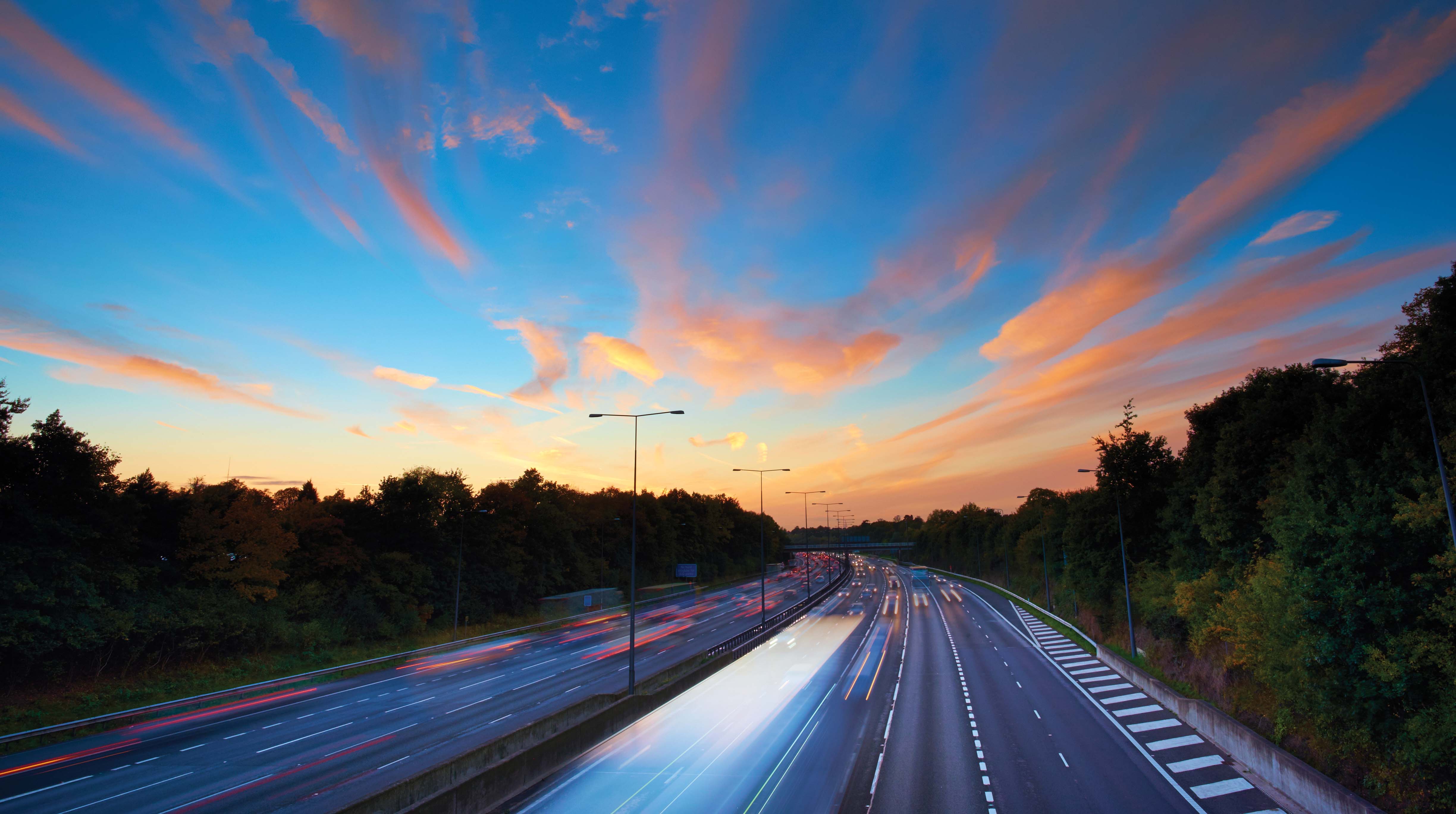 Dusk over a busy highway with light streaks from cars, framed by lush trees beneath a colourful sky filled with wispy clouds.