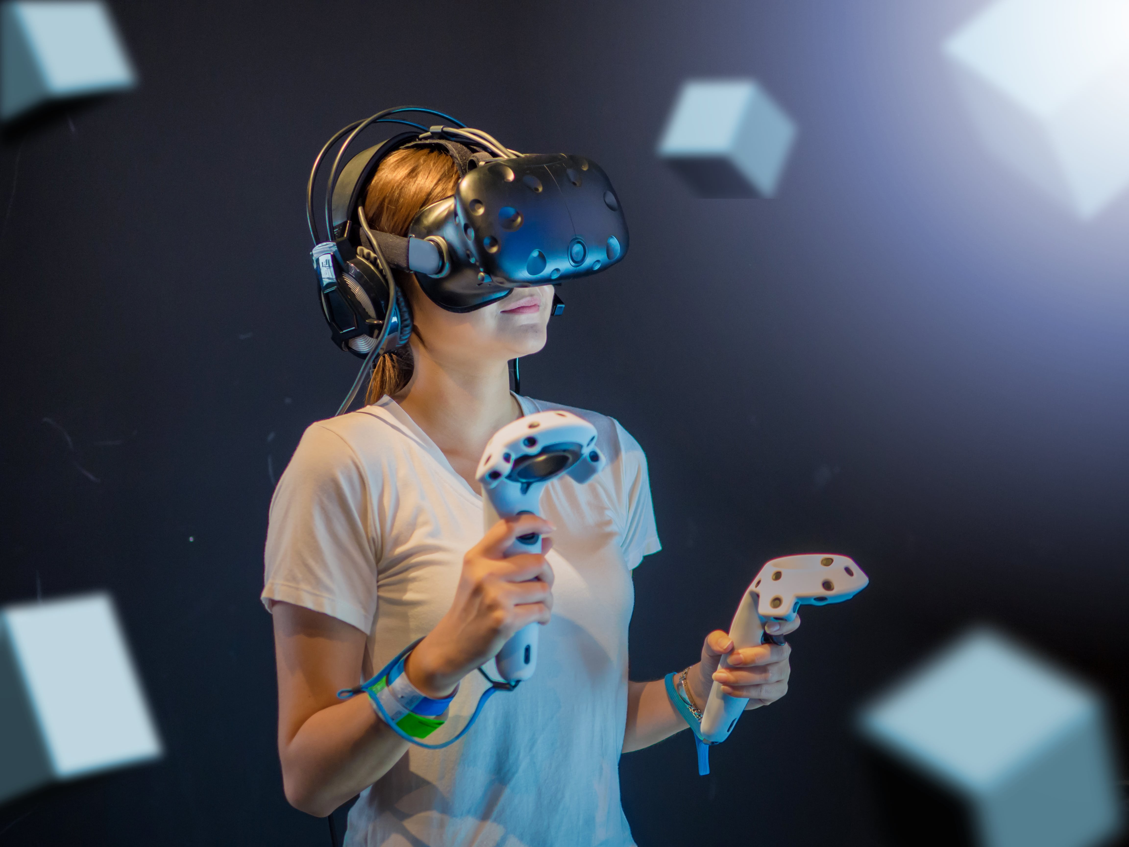 A woman wearing a VR headset and holding controllers, immersed in a virtual reality experience, with floating graphic cubes around her.