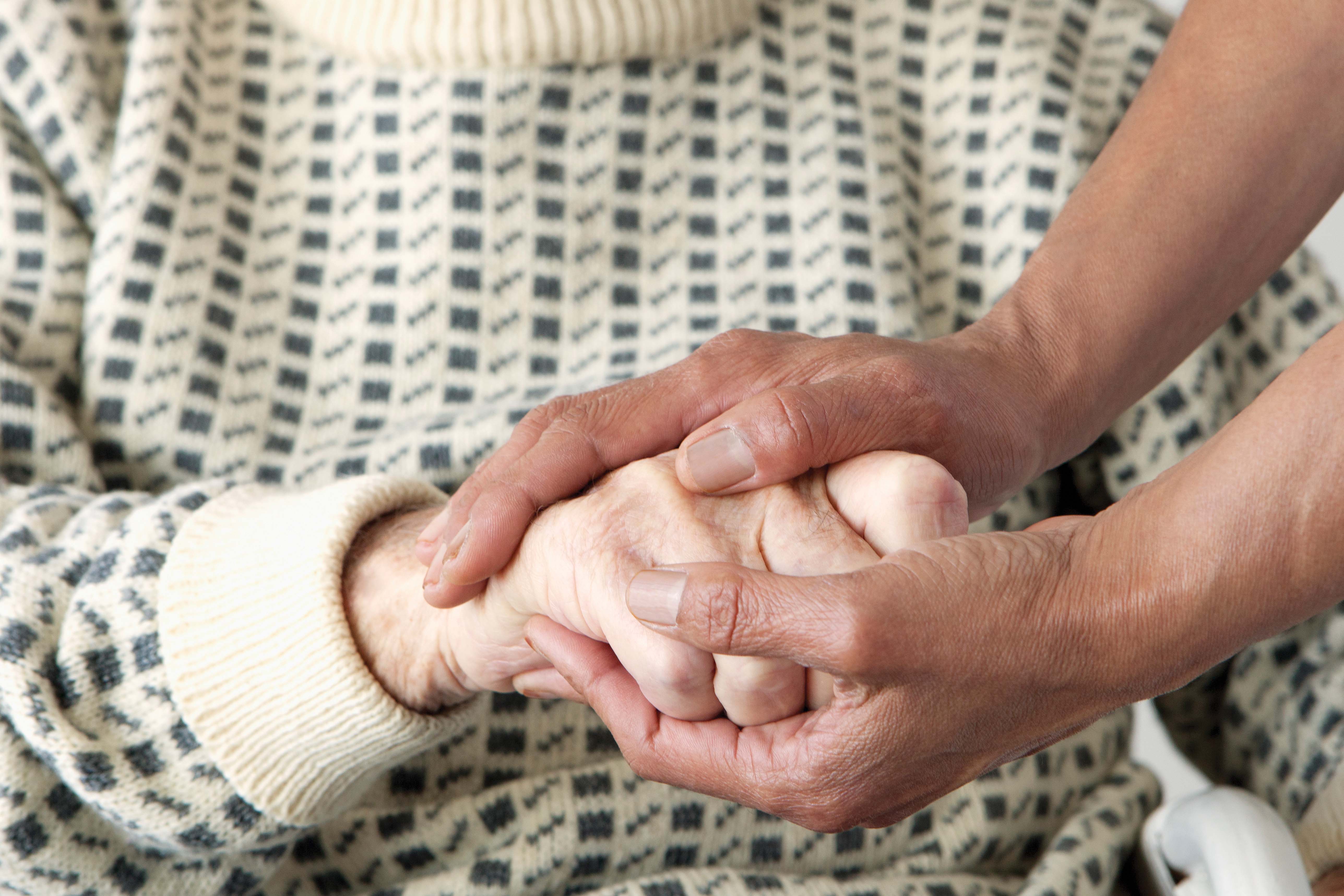 A caregiver gently holds the hand of an elderly person. Both hands are intertwined.