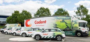 A woman wearing a denim jacket stands in a golden wheat field, raising her hand toward a colourful sunset sky.