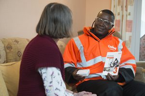 A Cadent engineer in an orange jacket sitting on a sofa with someone in discussion whilst holding a pamphlet