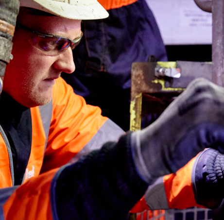 A worker in an orange safety jacket and helmet inspects machinery closely. He wears safety glasses and gloves, concentrating on his task in an industrial setting.