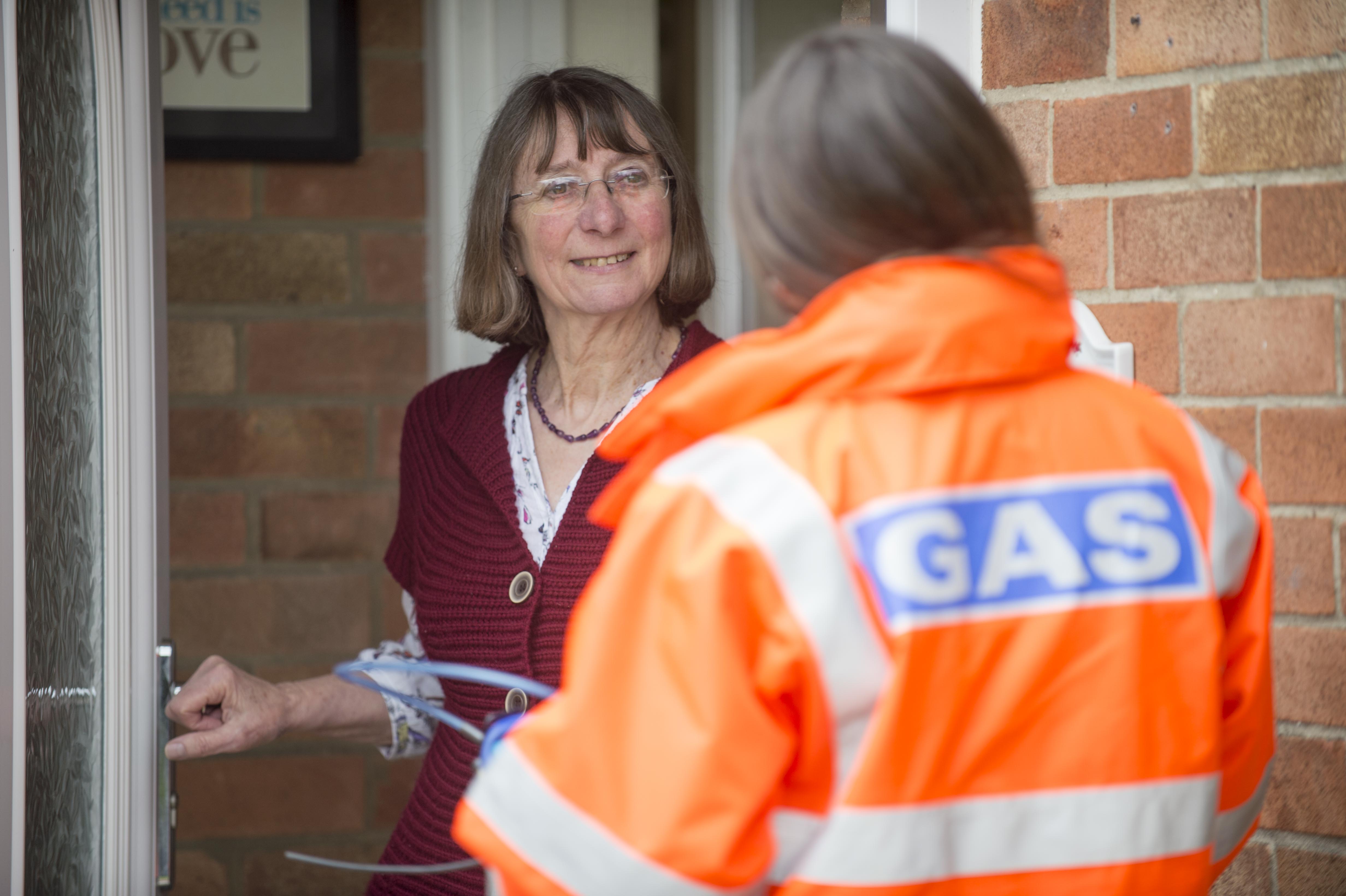 A gas worker in an orange uniform stands at the door, speaking to a woman in a cozy cardigan, both engaged in discussion.