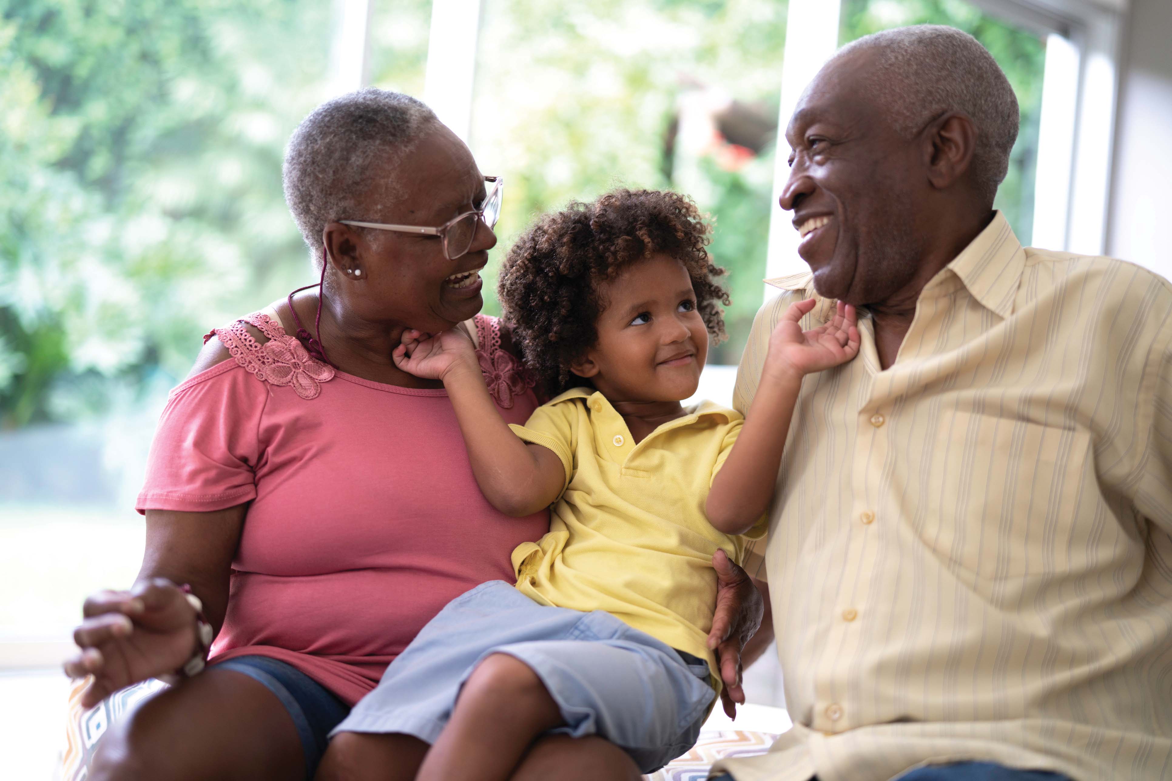 A child sits between two adults, smiling, in a bright room with large windows, surrounded by greenery outside.