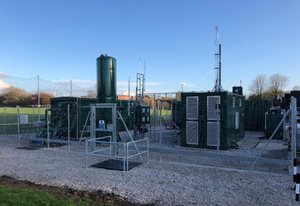 A green energy facility featuring storage tanks, generators, and communication towers, surrounded by a protective fence and open field.