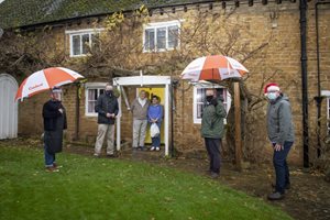 Five people wearing face masks stand under umbrellas at a cottage entrance, smiling, with autumn leaves in the background.