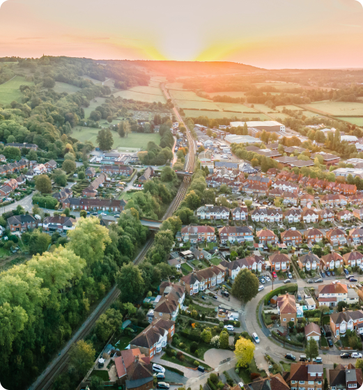 Aerial view of a residential suburban area with houses, roads, and greenery, under a sunset sky. A railway track cuts across the neighbourhood.