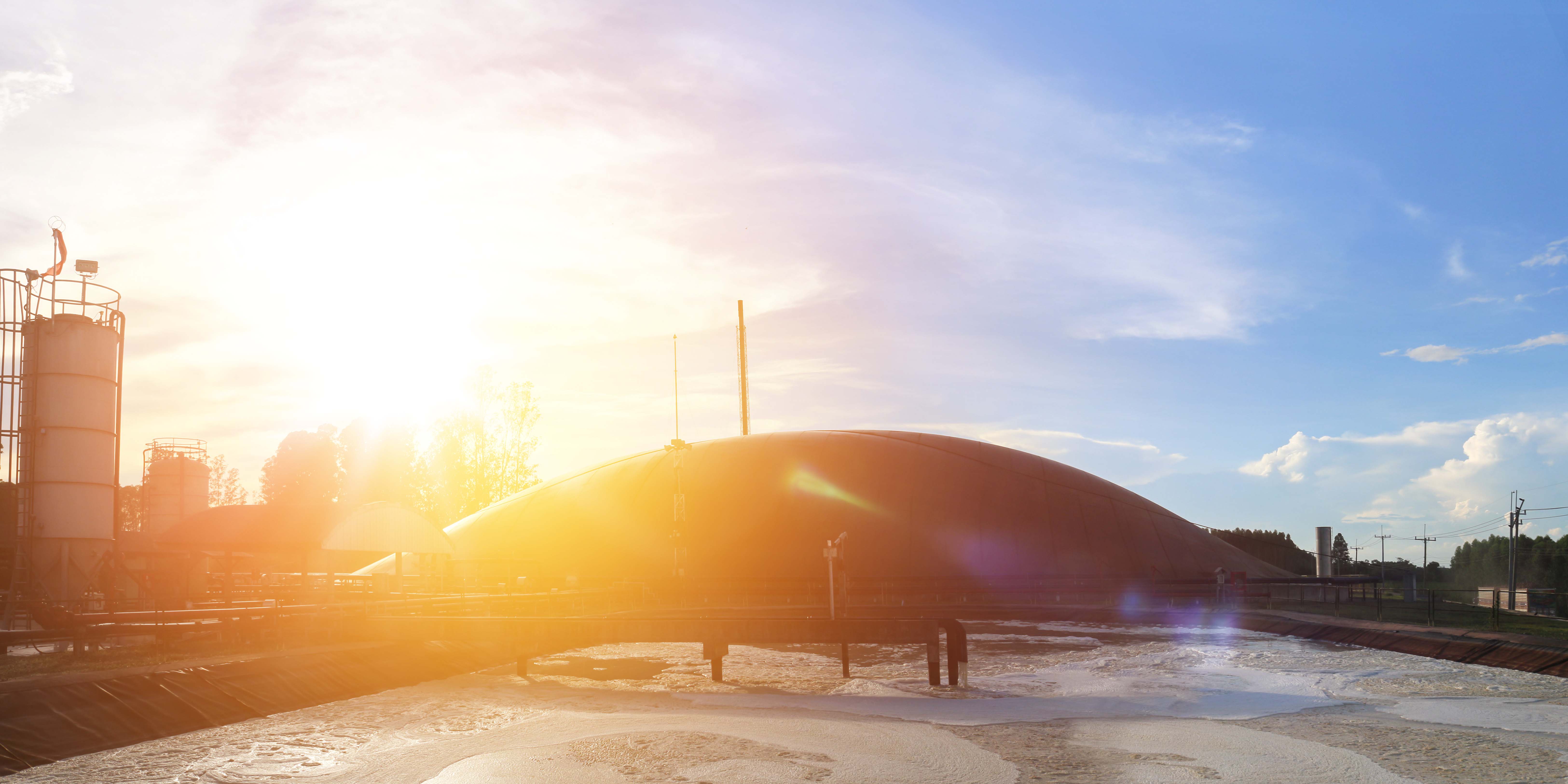 Sunset over a wastewater treatment facility with tanks and a large dome, reflecting warm light on bubbling water surfaces.